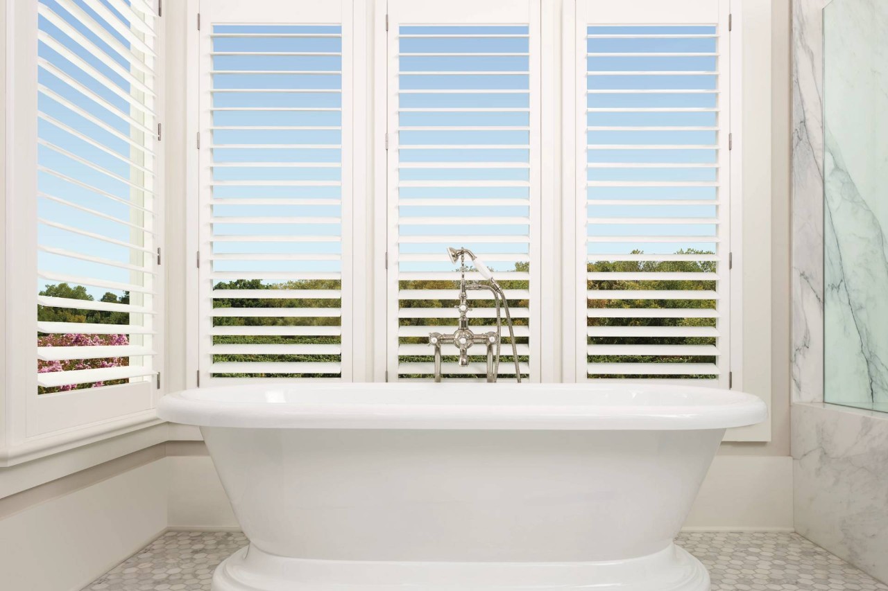 Open Hunter Douglas Palm Beach™ Polysatin™ Vinyl Shutters in a white bathroom with a stand-alone tub near Ocean City, MD